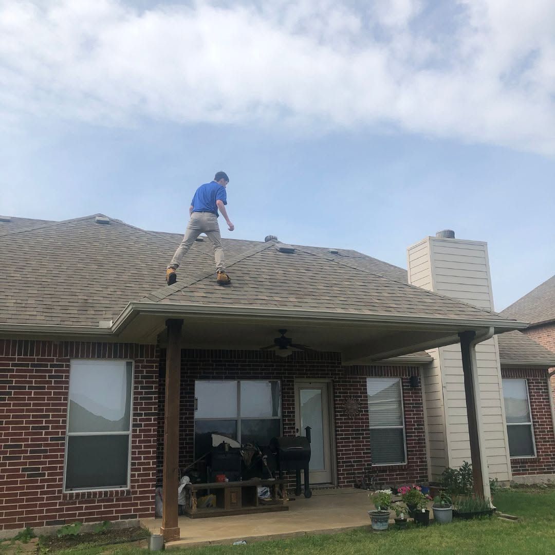 Person on a roof repairing shingles above a brick house with a covered porch