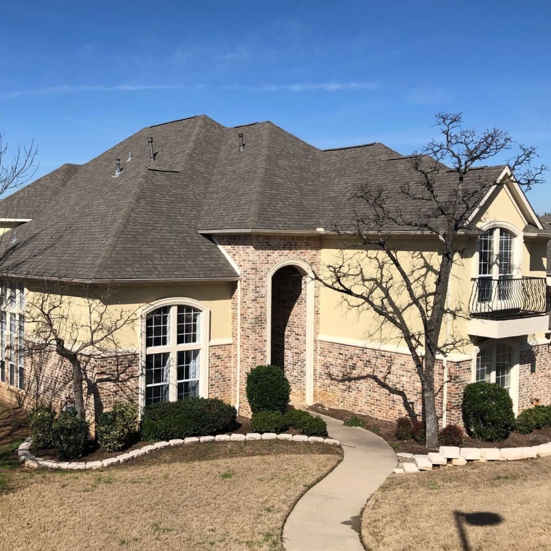 Two-story beige house with a dark roof, arched entry, and curved walkway under a clear blue sky