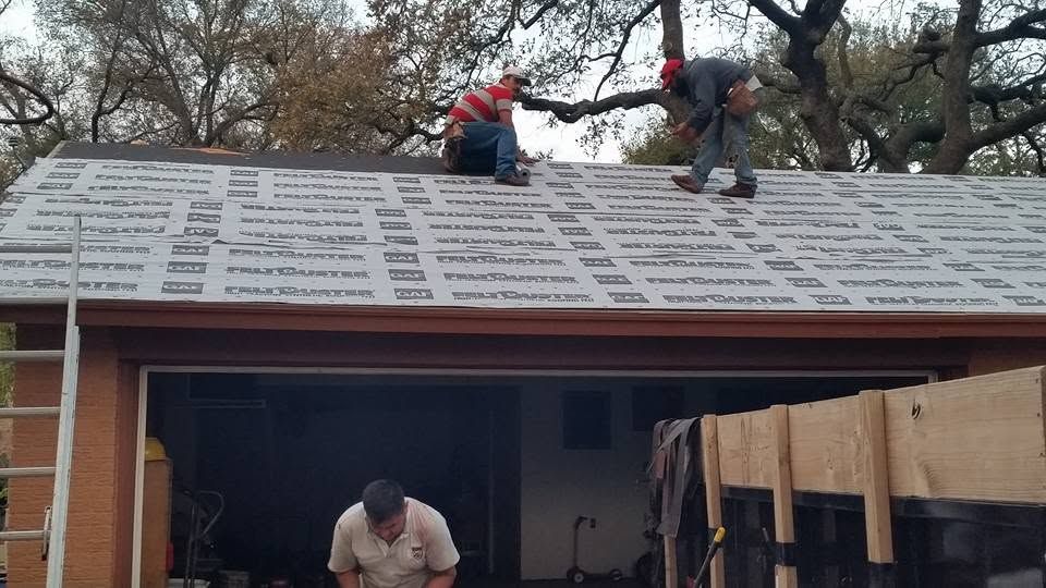 Workers installing shingles on a roof above a garage, with trees in the background.