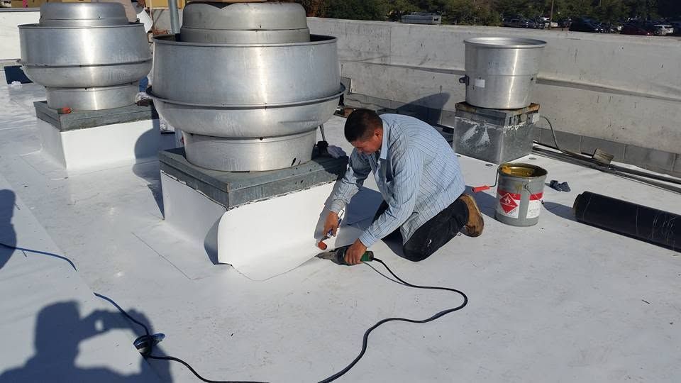 Worker on a rooftop repairing a white vent base beside large metal ventilation units.