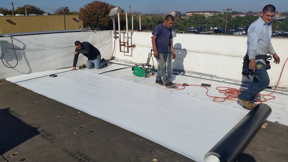 Workers installing a white rooftop membrane on a flat roof with tools and rolled material.