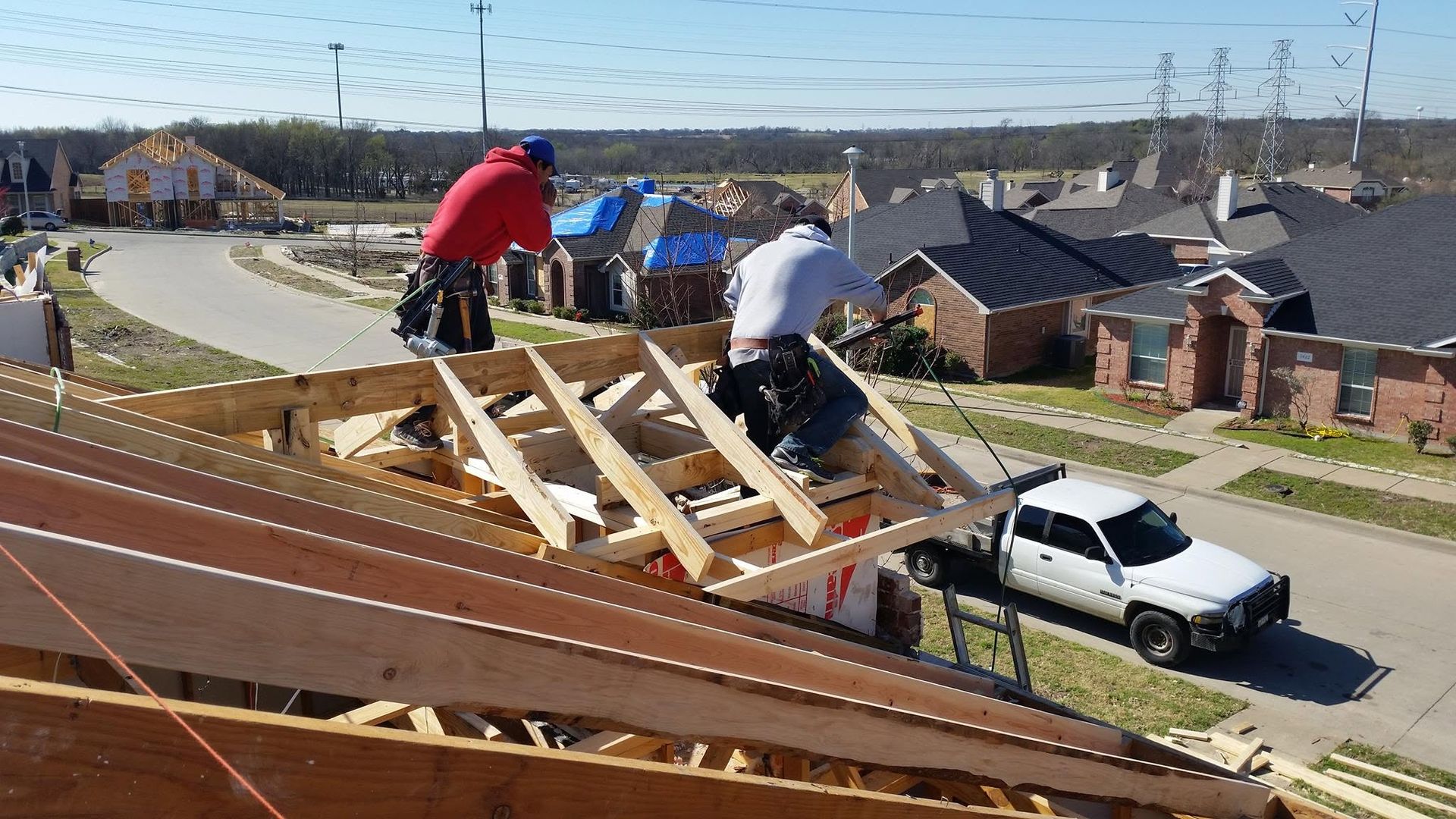 Two workers repairing a roof with wooden framing beside a white pickup in a suburban neighborhood