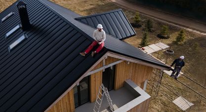 Two people installing roofing on a modern house, one on the roof and one on a ladder below.