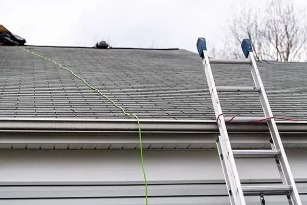 Two ladders lean against a gray shingled roof with a green rope hanging down.