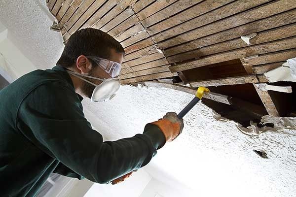 Worker wearing a mask and goggles uses a hammer to repair a damaged ceiling.