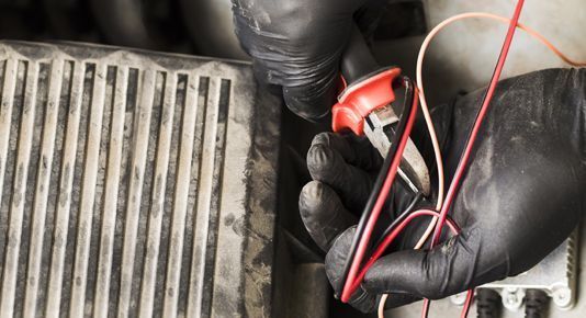 Gloved hands holding red and black wires near a car radiator or cooling component