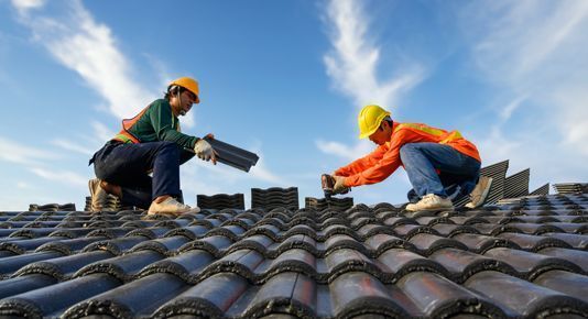 Two roofers in hard hats installing tiles on a sloped roof under a blue sky