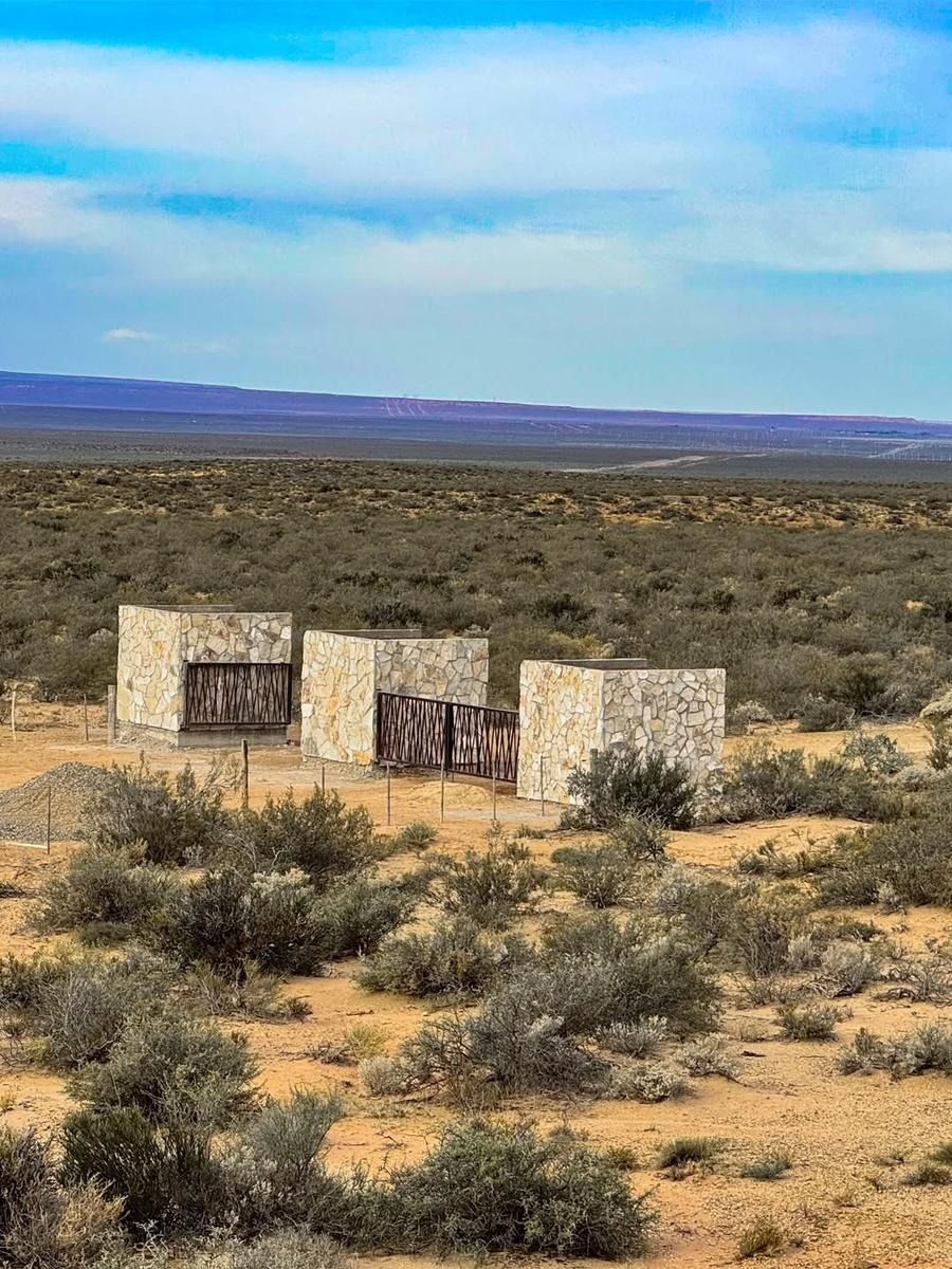 Estructura de piedra con tres pilares y una valla metálica en medio de un paisaje desértico arenoso bajo un cielo despejado.