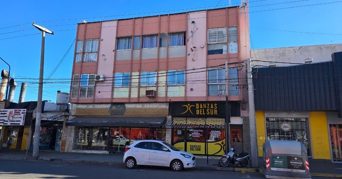 Vista de la calle de un edificio rosa desgastado con escaparates y coches aparcados bajo un cielo azul brillante.