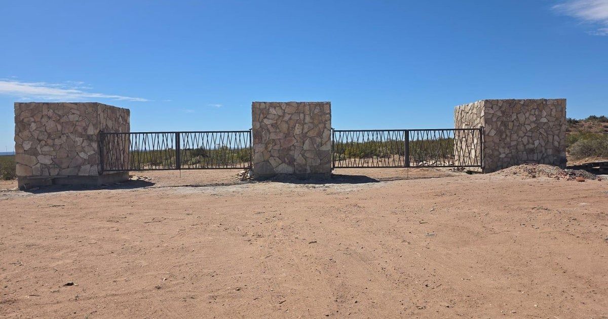 Estructura de piedra con tres pilares y una valla metálica en medio de un paisaje desértico arenoso bajo un cielo despejado.