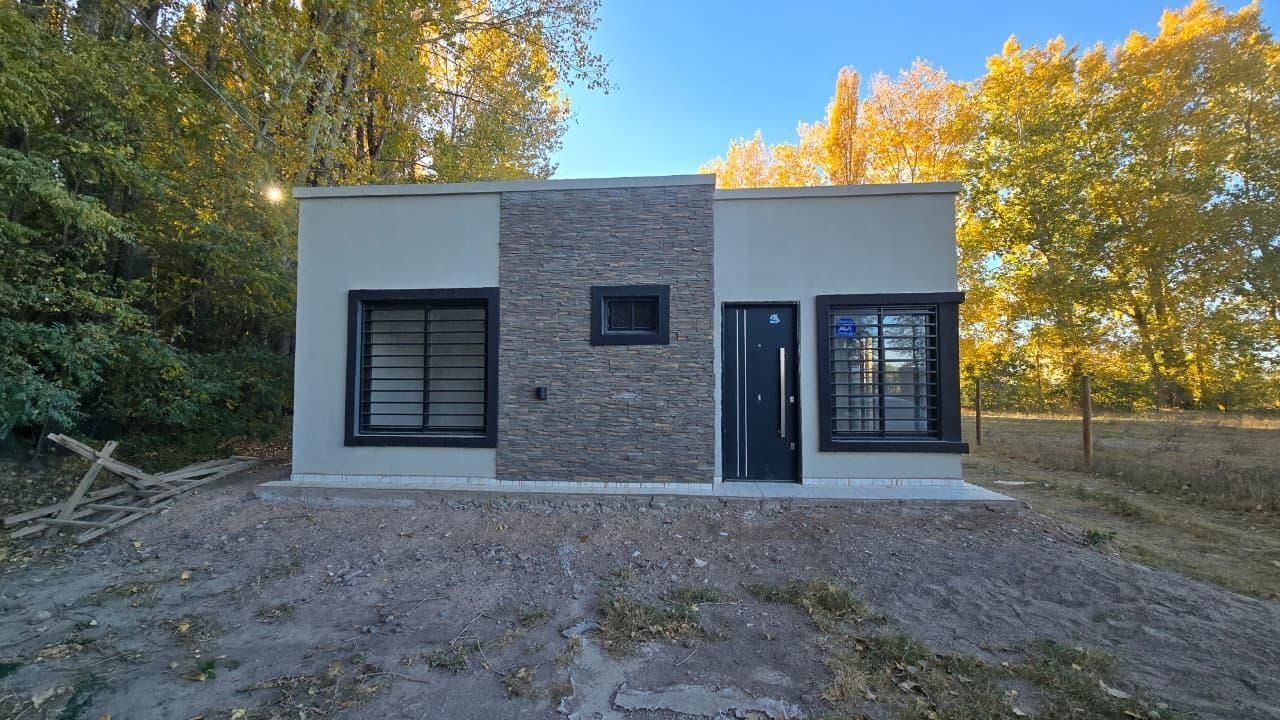 Pequeña casa moderna con fachada de piedra gris, ventanas negras y puerta principal en un terreno de tierra al atardecer.