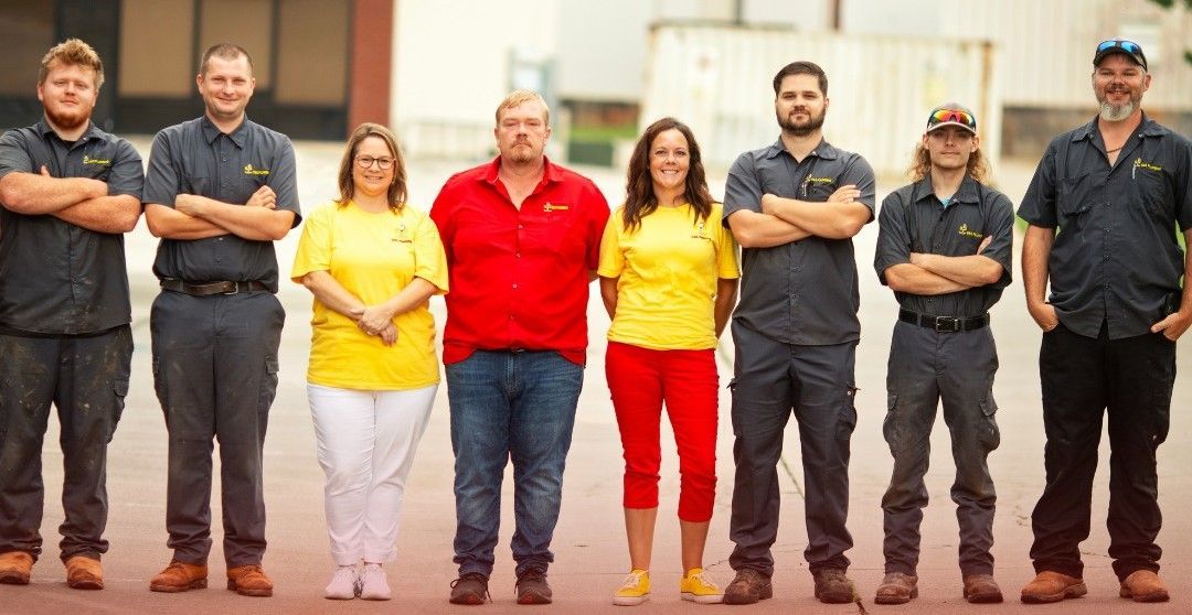 Group of eight people standing outdoors; some wearing yellow shirts, others in gray work shirts.