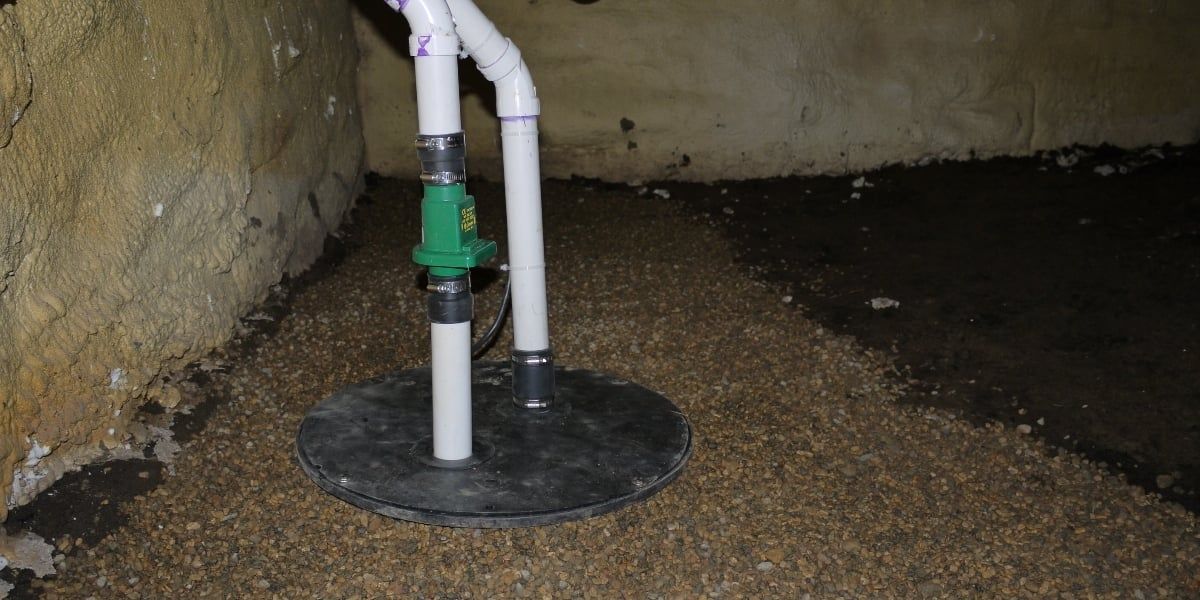A sump pump with white pipes and a green valve sits in a basement corner. The floor is covered in small pebbles.