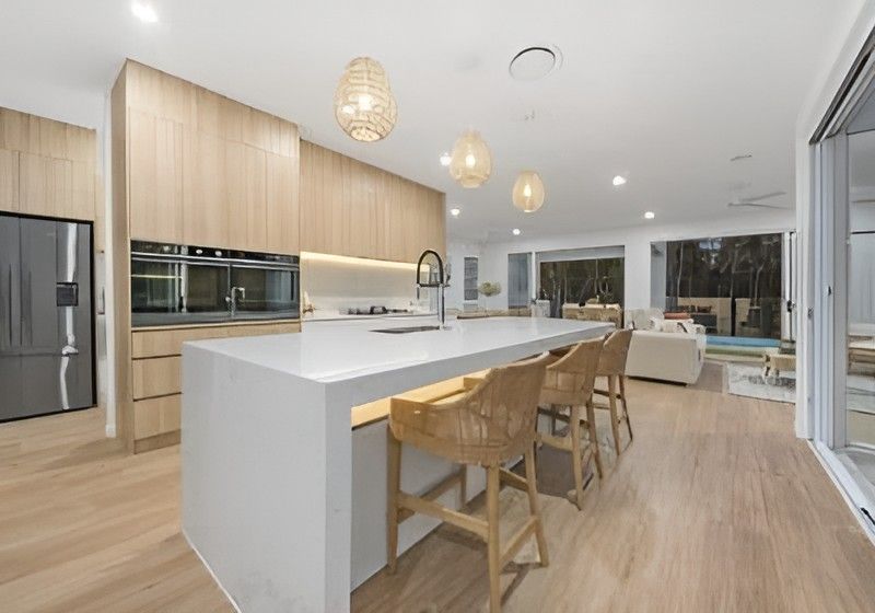 Modern Open Plan Kitchen with Light Wood Cabinetry, White Island, Woven Stools, and Pendant Lights — Totally Flooring Gold Coast In Bundall, QLD