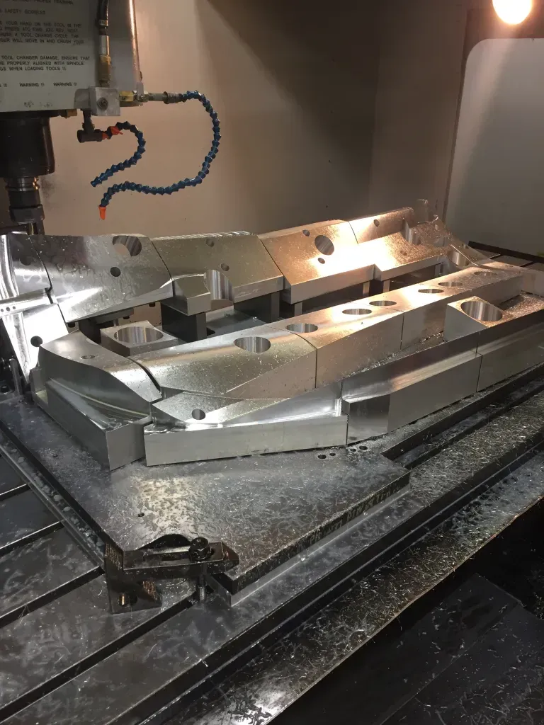 Metal CNC machine cutting a large aluminum fixture in a workshop, with coolant hoses and metal shavings visible.