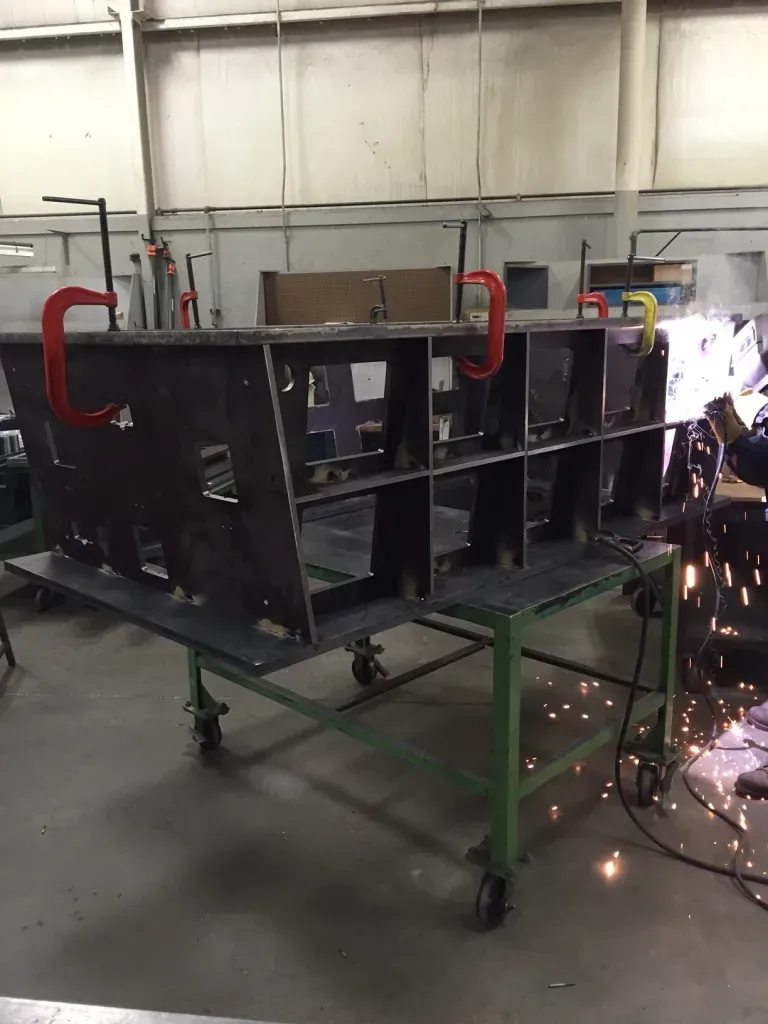 Worker welding a black metal frame on a green cart, with bright sparks in a workshop.