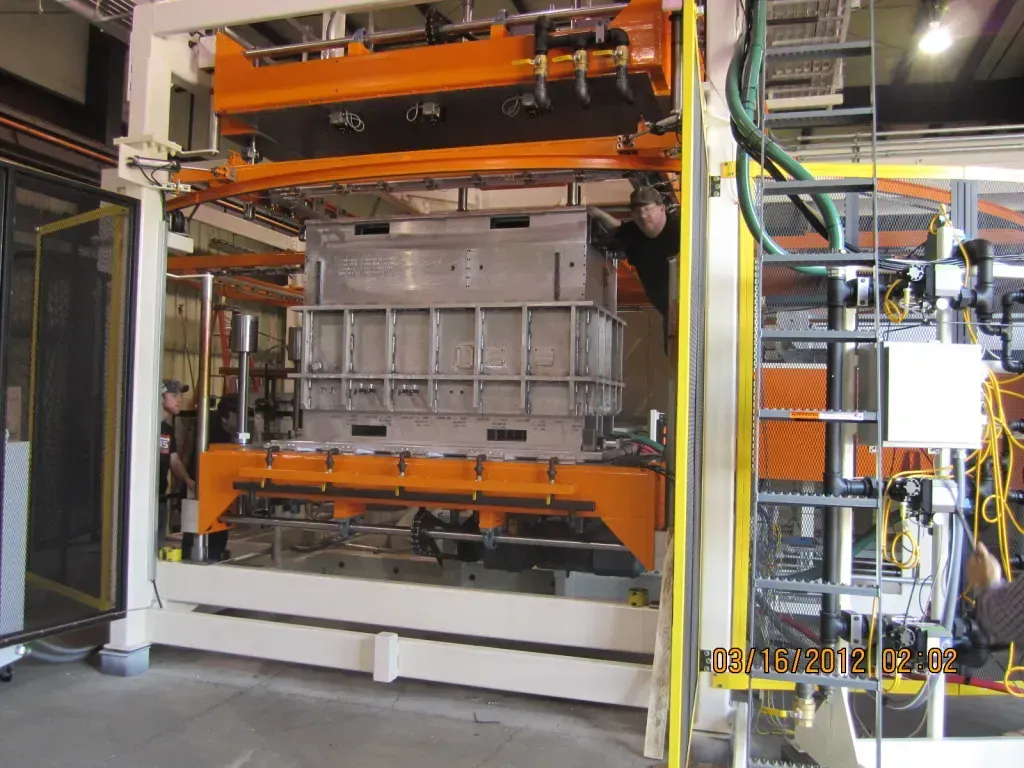 Industrial machine with orange overhead arms and a gray metal mold in a factory setting