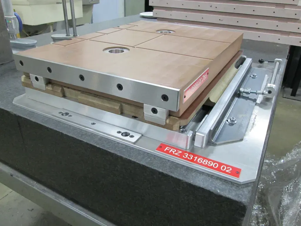 Metal machine fixture with a brown clamping plate mounted on a worktable in a workshop.