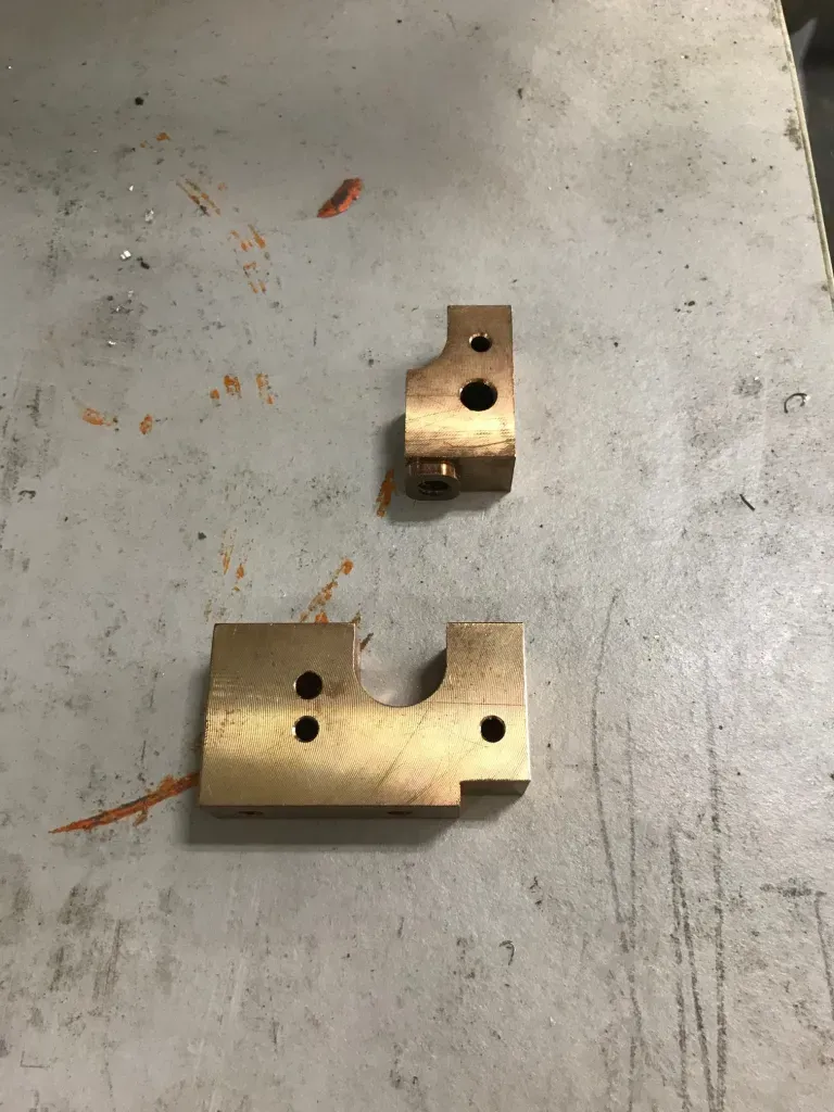 Two brass metal brackets on a gray concrete floor, one small and one larger.