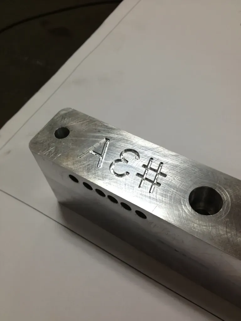 Metal block with engraved “4C#” and drilled holes on a white surface.