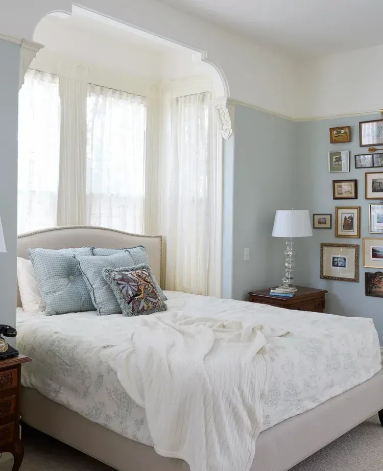 Bedroom with bed, bay window, light blue walls, and framed pictures.