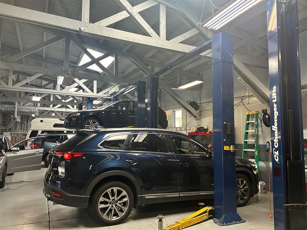 Dark SUV on a car lift inside an auto repair shop with other vehicles and blue support columns. | Marin Automotive