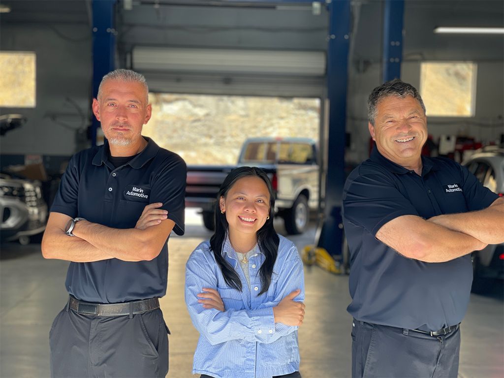 Three people in a garage, smiling with arms crossed, standing in front of a car lift. | Marin Automotive
