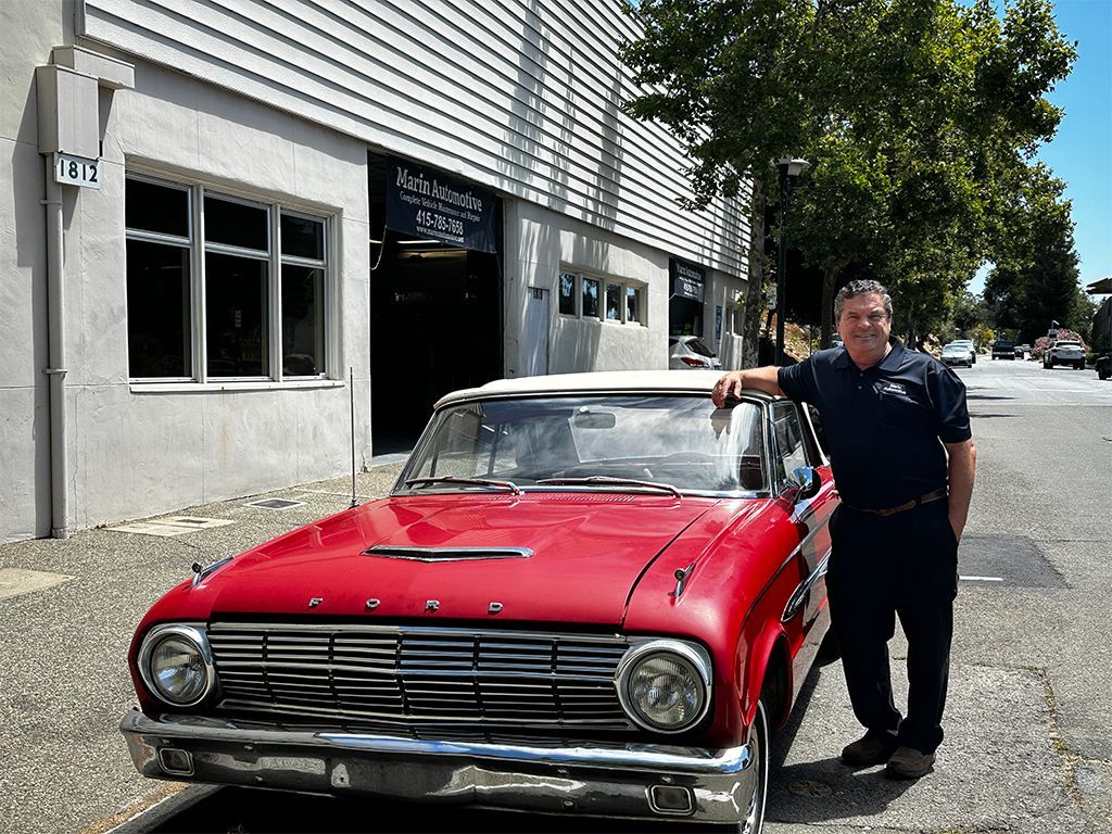 Man standing beside a red vintage car parked outside a building on a sunny street. | Marin Automotive