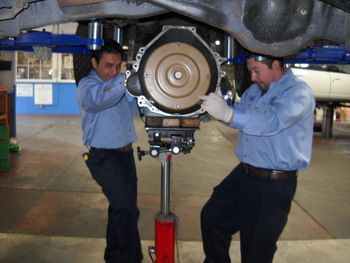 Two mechanics in blue shirts work beneath a raised car, supporting a transmission on a jack. | Marin Automotive