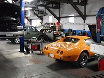 Classic cars in an auto shop, including a bright orange sports car and a silver car on lifts. | Marin Automotive