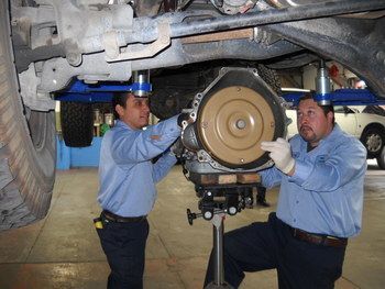 Two mechanics in blue uniforms work on a vehicle’s brake assembly under a raised car. | Marin Automotive