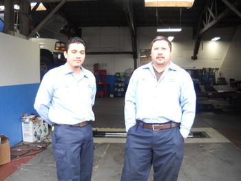 Two men standing in a garage, wearing light blue shirts and dark pants, facing the camera. | Marin Automotive