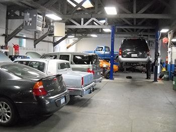 Auto repair shop with several parked cars and trucks inside a garage, including a lifted SUV on a hoist | Marin Automotive