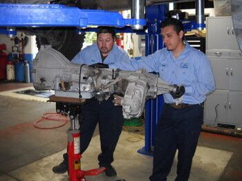 Two mechanics in blue shirts lifting a car transmission in a repair shop | Marin Automotive