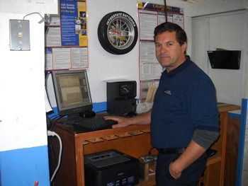 Man standing at a computer desk in a workshop, with tools and a dartboard on the wall. | Marin Automotive
