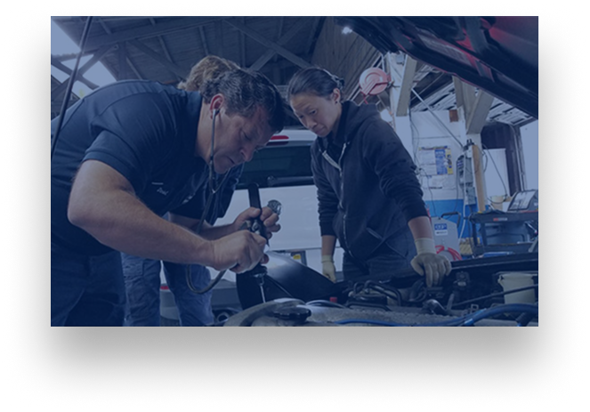 Two mechanics working under a blue car hood in a garage, inspecting engine parts.