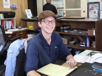 Smiling person wearing a cap and glasses sits at a desk in an office, writing on paper. | Marin Automotive