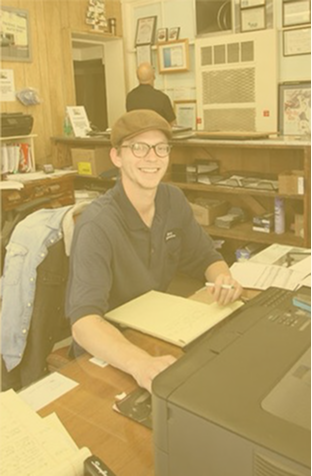 Smiling person in a black cap at a desk in a cluttered office, with a printer in the foreground.