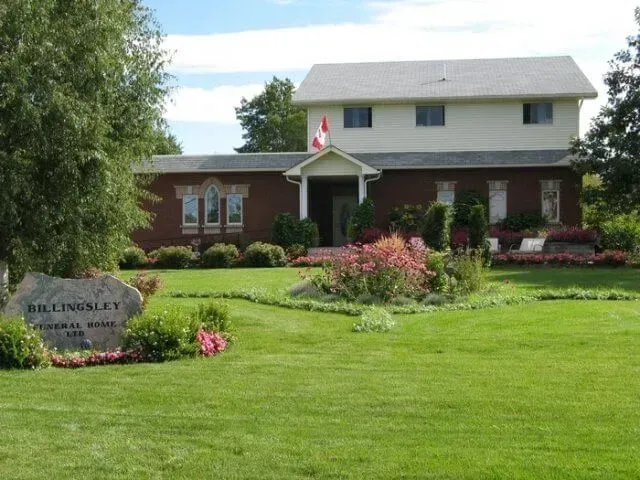 Brick building with white upper floor, flower garden, and a stone sign in front of a green lawn.