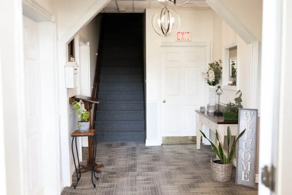 Bright entryway with dark staircase, white door, hanging lantern, and potted plants on patterned tile floor