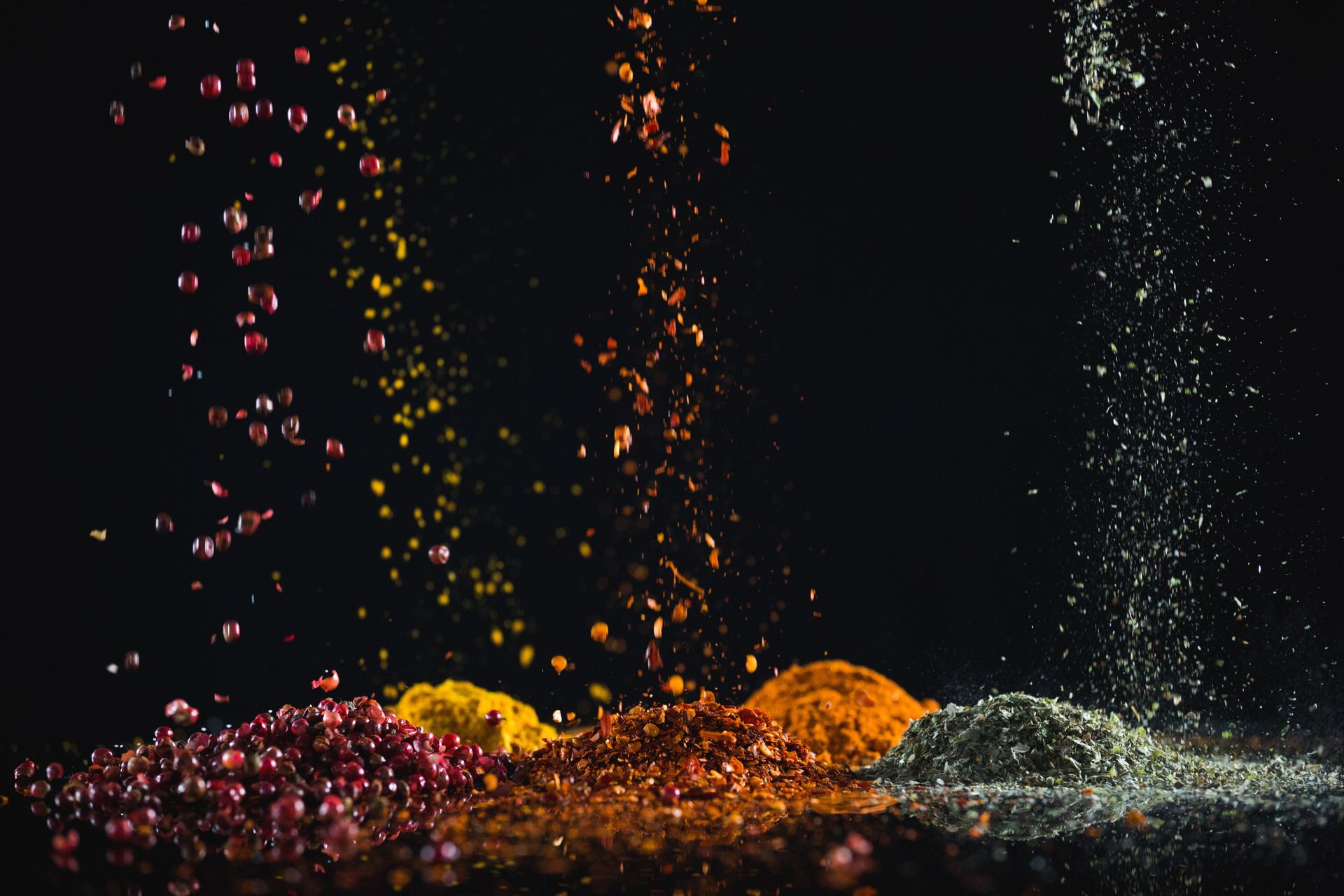 Colorful spices falling against a black background. Red, yellow, orange, and gray piles form below.