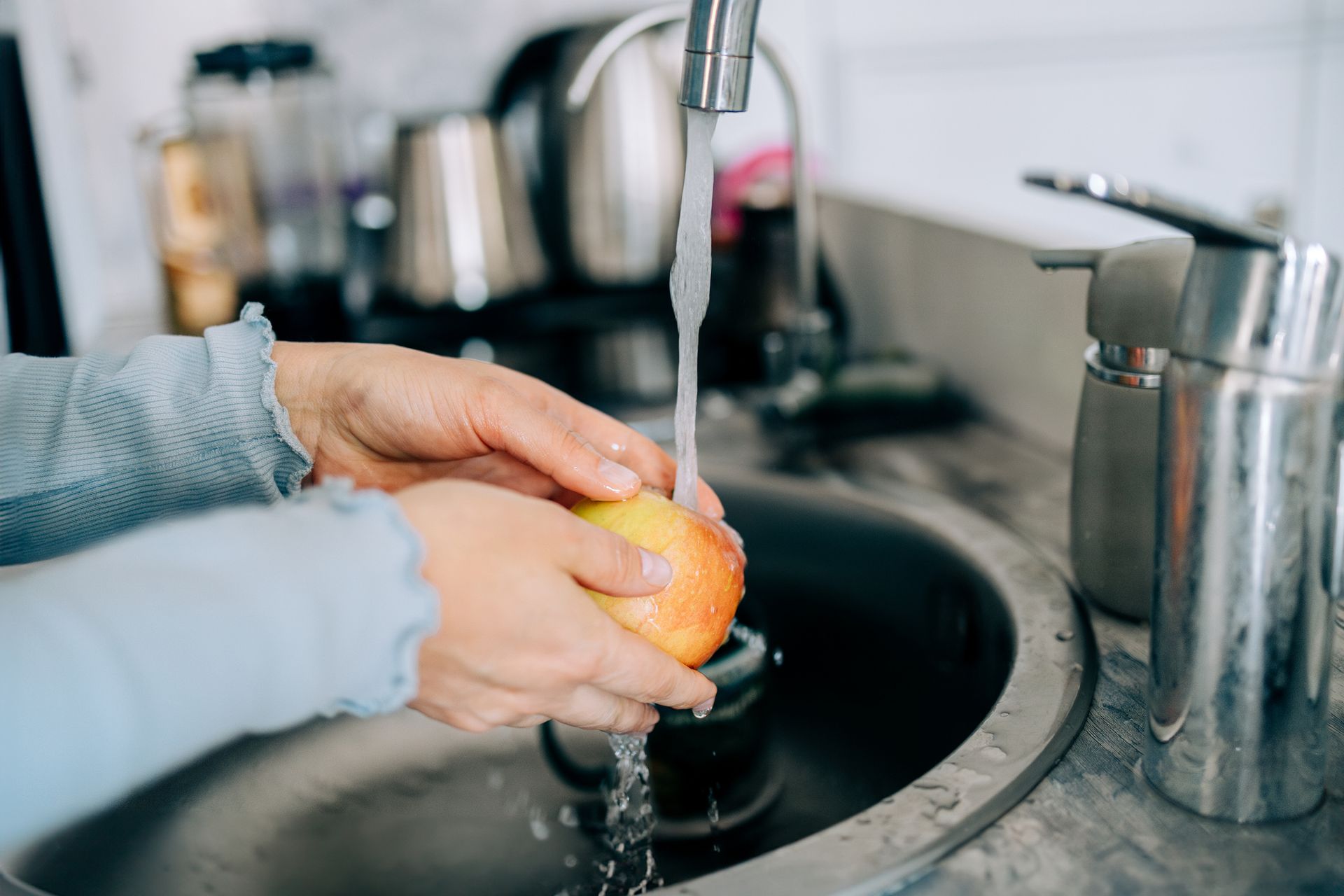 A close-up of hands washing a fresh apple under a kitchen sink faucet. A close-up of hands washing a fresh apple under a kitchen sink faucet.