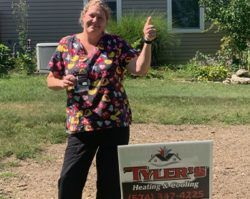 A woman is standing in front of a house holding a sign and giving a thumbs up.