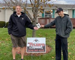 Two men are standing in front of a house holding a sign.