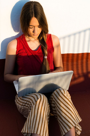 Teenager sitting, looking down at a laptop in warm sunlight, with striped pants and a red top
