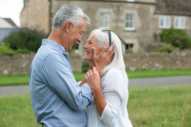 An elderly couple is dancing in a field in front of a house.