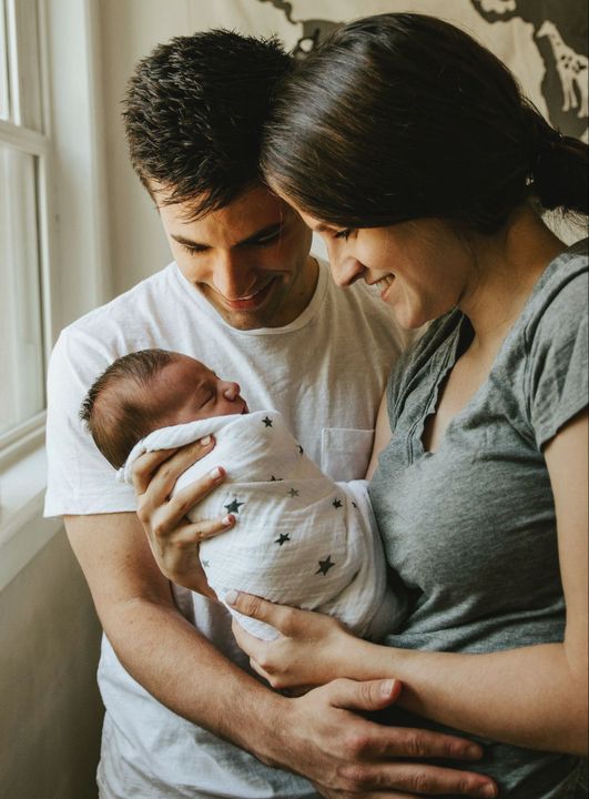 A man and a woman are holding a newborn baby in front of a window.