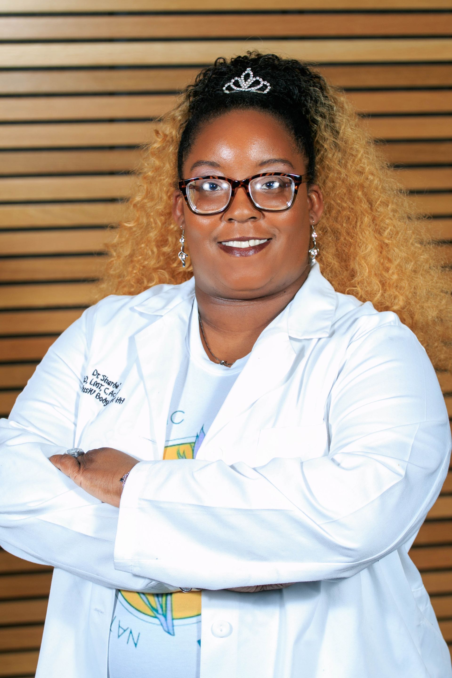 Smiling doctor in white coat with arms crossed, standing against a wooden slat wall.