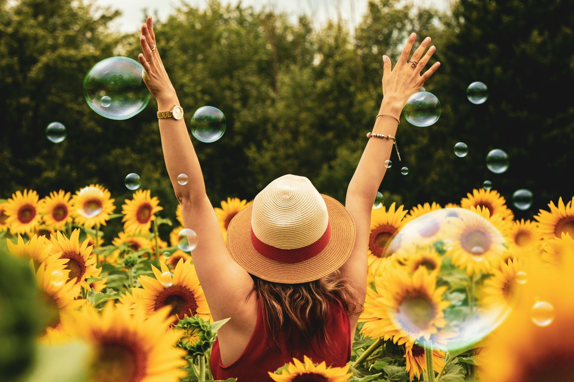 Person in a straw hat raises arms in a sunflower field with floating soap bubbles.