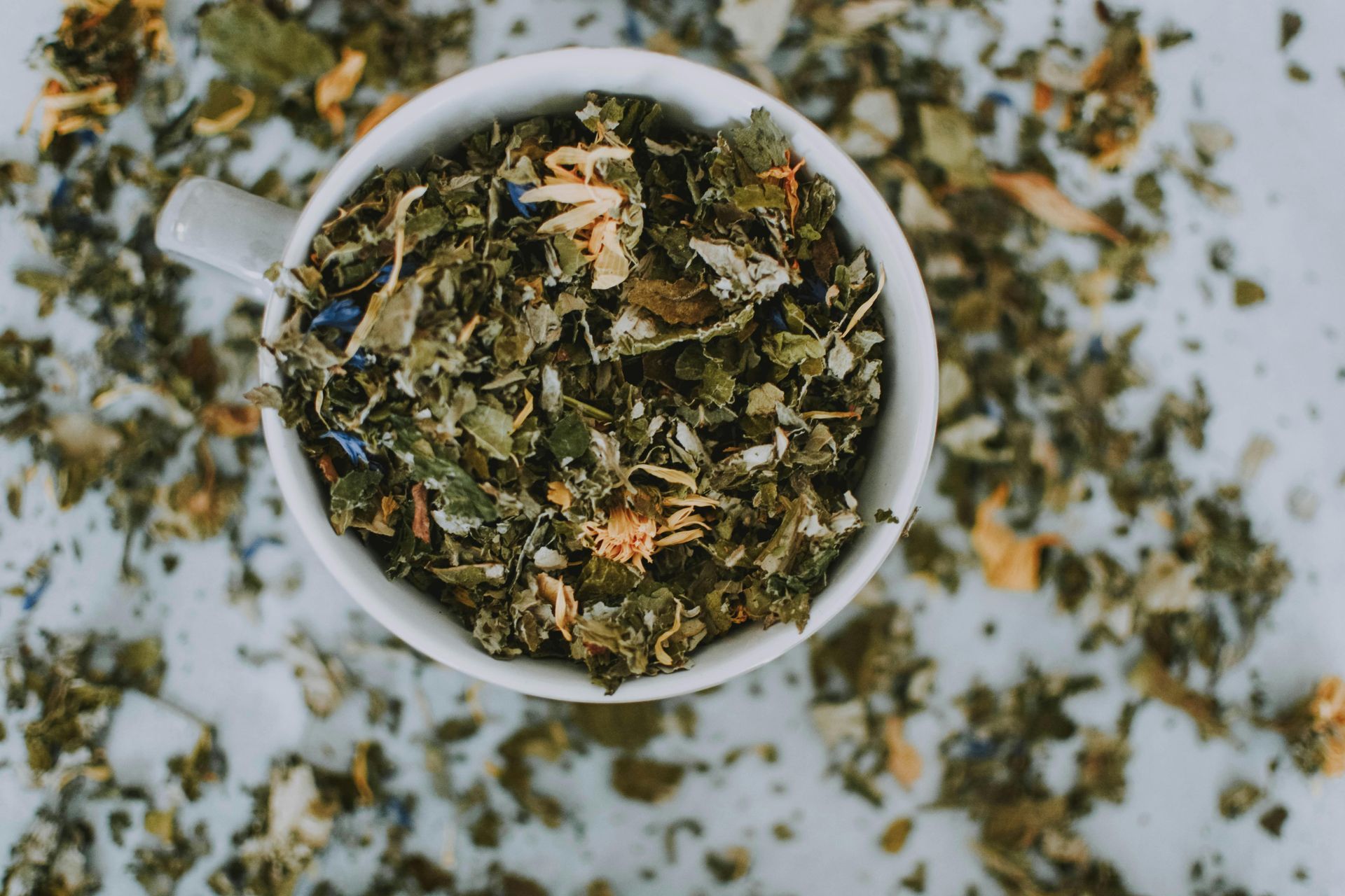 White cup of loose dried herbs and flowers on a speckled white surface